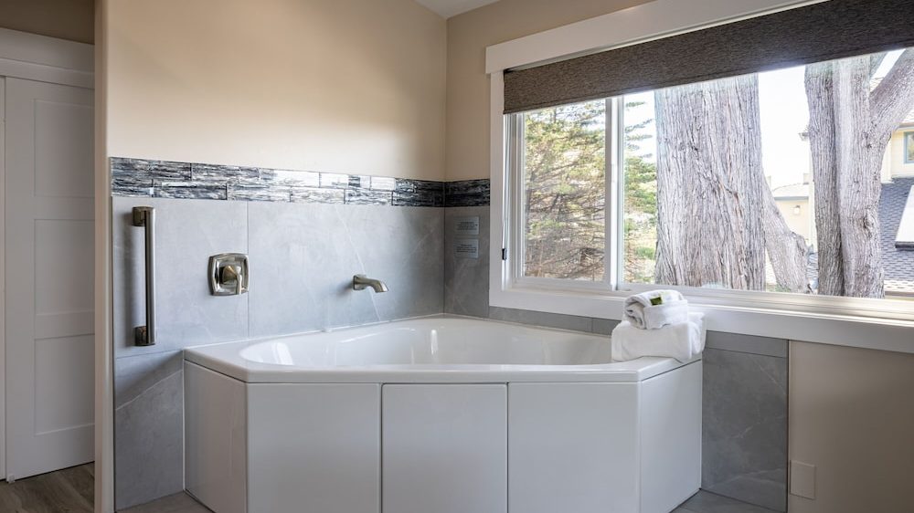 Corner bathtub with white panels and silver fixtures, set against gray tiled walls with a decorative black and gray tile border, next to a large window with a dark roller shade and a stack of white towels on the tub edge.
