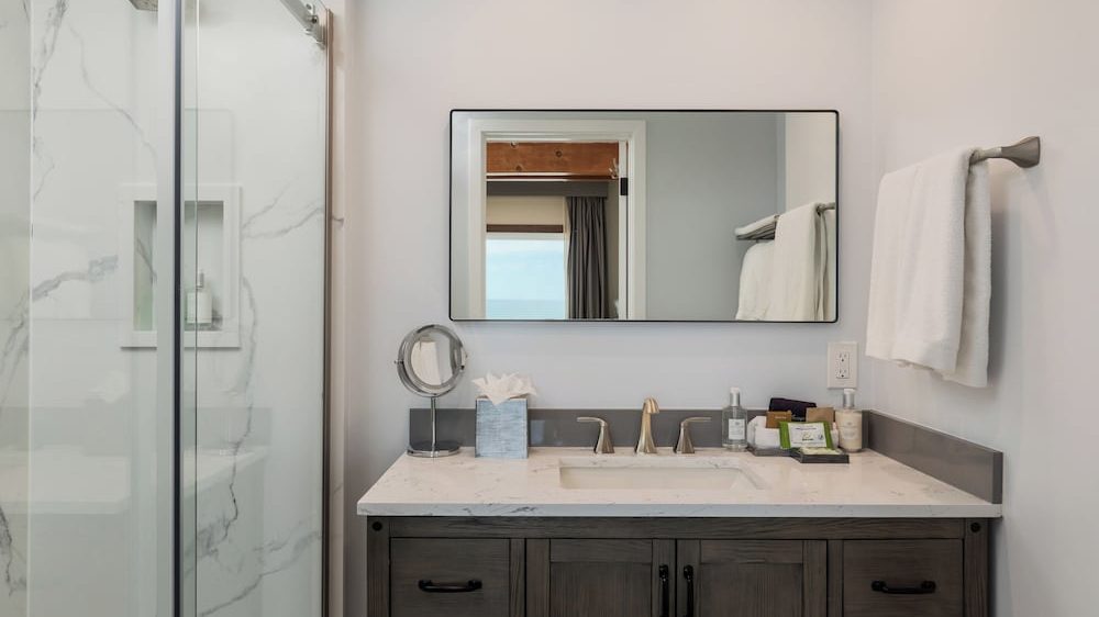 Bathroom vanity with a white marble countertop, built-in sink, and brushed nickel faucet. A rectangular mirror with a thin black frame hangs above the sink. On the countertop are a tissue box, a small round makeup mirror, and various toiletries. A white towel hangs on a metal towel bar mounted on the wall to the right. To the left, a glass shower door reveals a shower with white marble-patterned tiles. The vanity cabinet is dark wood with black handles.