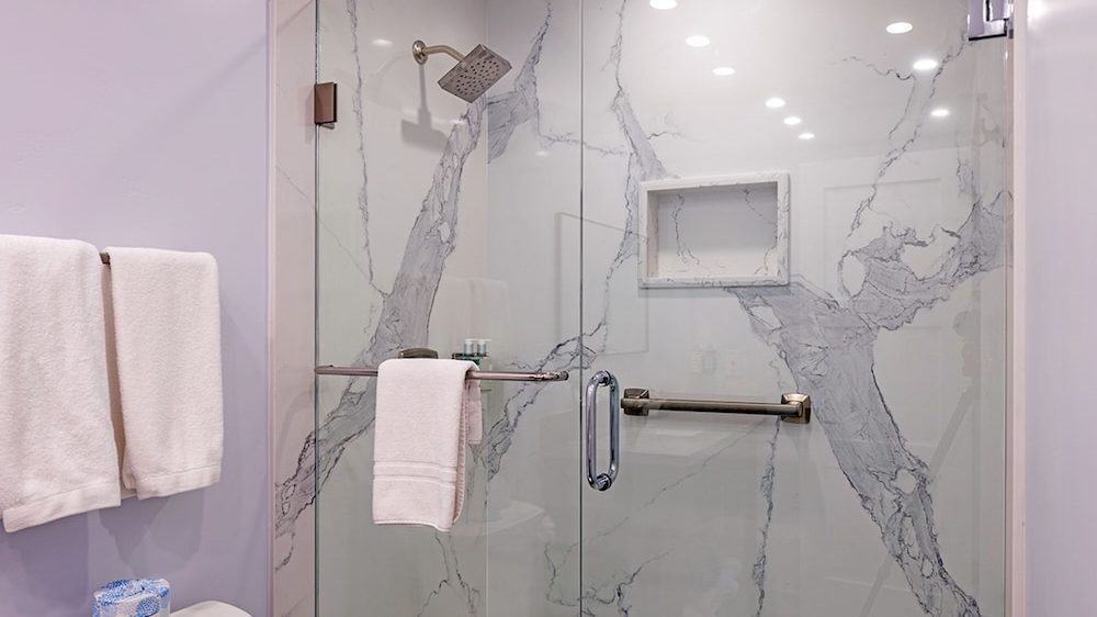 Glass-enclosed shower with white marble walls featuring gray veining, a square rain showerhead, and a built-in niche. White towels hang on a bar inside the shower and on a towel rack outside. A roll of Cottonelle toilet paper is visible on top of a toilet tank.
