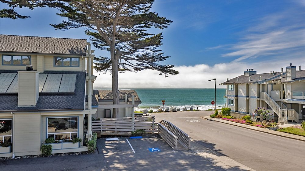 Two light-colored residential buildings with gray roofs are situated near a coastal road leading to the ocean. A large tree stands between the buildings, and the ocean waves are visible in the background under a partly cloudy blue sky. There is a parking area with a handicap space in the foreground.