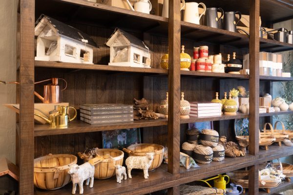 Wooden shelves filled with various decorative and gardening items including white birdhouse models, metal watering cans in copper, gold, and black, balls of twine in different colors, stacks of books titled "Private Gardens of Santa Barbara," wooden baskets, small white sheep figurines, dried flowers, and other small decorative objects.