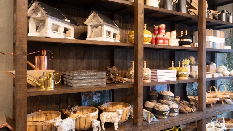 Wooden shelves filled with various decorative and gardening items including white birdhouse models, metal watering cans in copper, gold, and black, balls of twine in different colors, stacks of books titled "Private Gardens of Santa Barbara," wooden baskets, small white sheep figurines, dried flowers, and other small decorative objects.