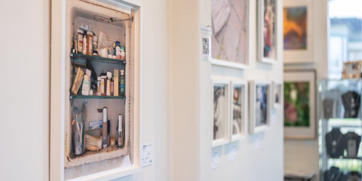 Framed artwork of a medicine cabinet filled with various bottles, tubes, and containers, mounted on a white gallery wall. Other framed artworks are visible in the background, slightly out of focus.