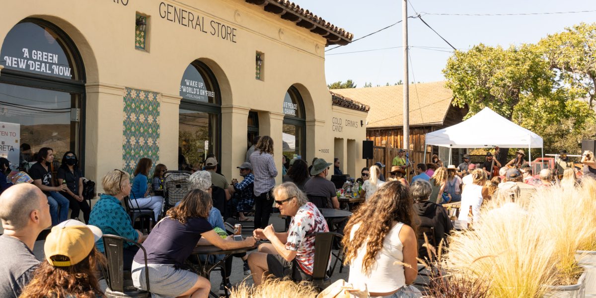 Outdoor gathering at San Gregorio General Store with people seated at tables and on chairs, facing a white canopy tent where a live band is performing. The building has a beige stucco exterior with arched windows and decorative tiles. Potted ornamental grasses line the foreground. The scene is sunny with clear skies.