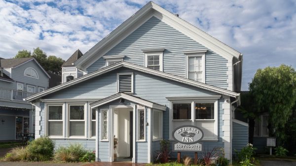 A two-story light blue wooden building with white trim, featuring a peaked roof and multiple windows. The entrance has a small covered porch with decorative trim. In front of the building is a sign that reads "BY THE SEA INN" with a phone number and website below. There are plants and shrubs along the front of the building. The sky is partly cloudy.