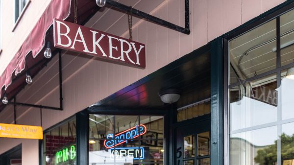 Storefront with a hanging sign reading “BAKERY” above a window with neon “OPEN” and “Sandwiches” signs.
