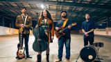 Four musicians stand in a spacious, industrial indoor setting. From left to right: a man in a brown jacket and cap holding an electric bass guitar, a woman with long curly hair wearing a brown coat and boots holding a cello, a man with glasses and a cap wearing a dark shirt and jeans playing an electric guitar, and a man in a dark shirt standing behind a bass drum and snare drum. The lighting is warm and the ceiling has exposed beams and panels.
