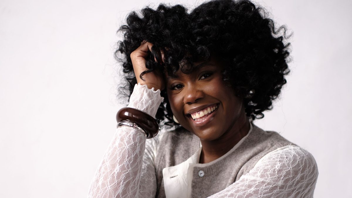 A smiling woman with curly black hair wearing a white lace long-sleeve top under a gray vest. She has large dark bangles on her wrist and is resting her head on her hand against a plain light background.