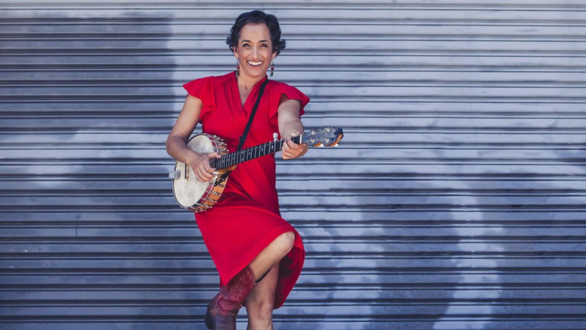 A woman wearing a red dress and brown boots is playing a banjo in front of a gray metal shutter. She is smiling and has one leg lifted.