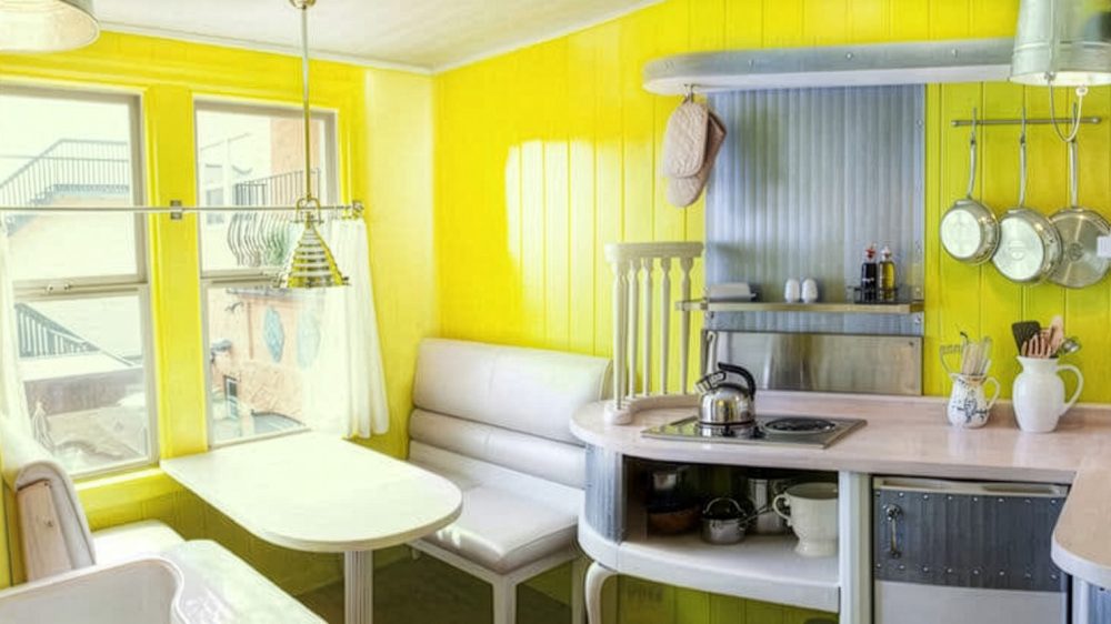 A small kitchen corner with bright yellow walls, featuring a white cushioned bench seating around a white table by two large windows with sheer white curtains. The kitchen counter is light-colored with a built-in stovetop and a silver kettle on it. Above the counter, there is a metal backsplash with hanging pots and pans, and two oven mitts hanging from a rail. Utensils are stored in white ceramic pitchers on the counter. A pendant light with a metallic shade hangs over the table.