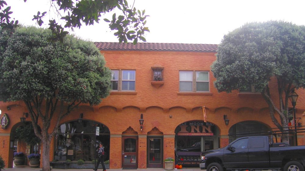 Two large, leafy trees flank the entrance of a two-story building with textured orange walls and a red-tiled roof. The building has multiple windows with shutters on the upper floor and arched doorways on the ground floor. A black pickup truck is parked in front on the right side, and a person is walking near the center entrance. There are potted plants and a sign indicating "Half Moon Bay Inn" visible near the entrance.
