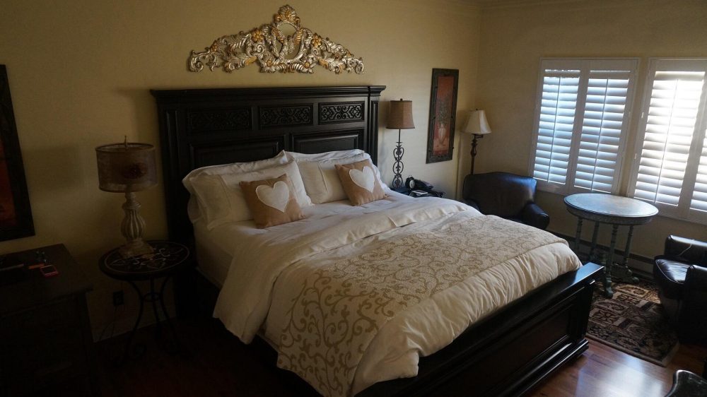 King-sized bed with a dark wooden headboard and footboard, dressed in white bedding and a beige patterned throw. Two beige pillows with white heart designs are placed on the bed. The room features two side tables with lamps, a small round table near the window, two dark leather armchairs, and a decorative wall piece above the headboard. Light filters through white plantation shutters on the windows. The floor is wooden with a small patterned rug near the seating area.