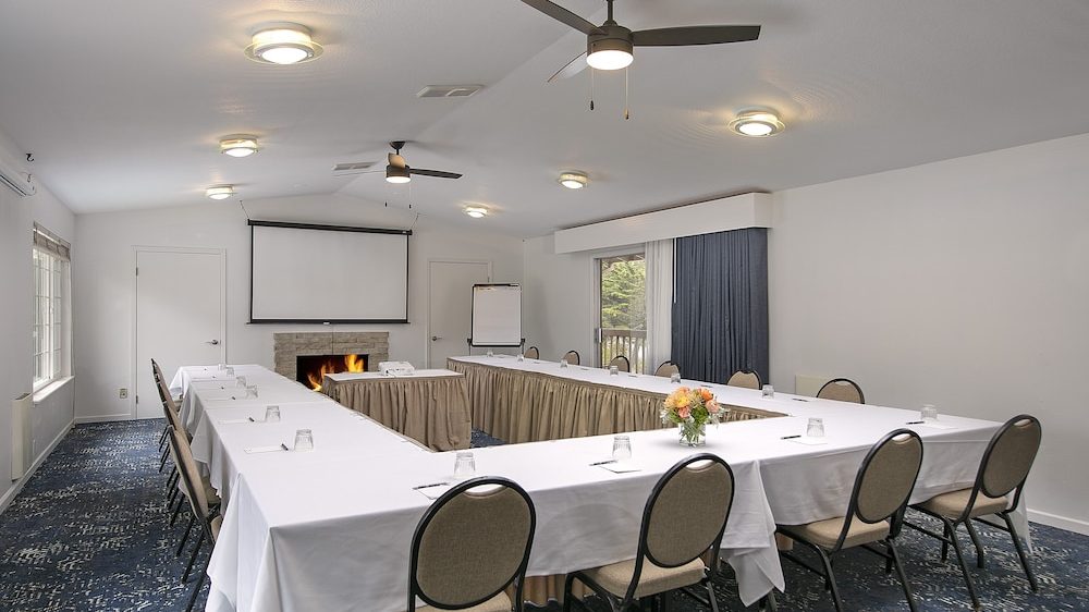 Conference room with U-shaped tables covered in white tablecloths, beige chairs, a flower arrangement in the center, a fireplace with a fire burning, a projection screen above the fireplace, a flip chart, ceiling fans, and blue patterned carpet.