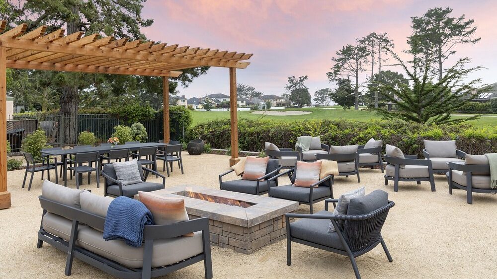 Outdoor patio area with modern gray cushioned seating arranged around a rectangular stone fire pit. A wooden pergola covers a dining table with chairs in the background. The setting overlooks a green lawn and trees under a pink and purple sunset sky.