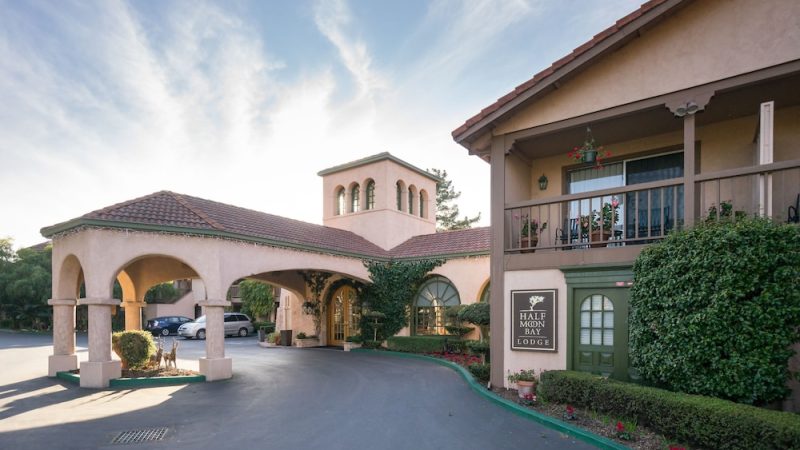 A Mediterranean-style building with a tiled roof and arched entranceway, featuring a sign that reads "Half Moon Bay Lodge." The structure has a covered driveway, green doors, and a balcony with potted plants. Ivy and shrubs decorate the exterior, and several cars are parked in the background under a partly cloudy sky.