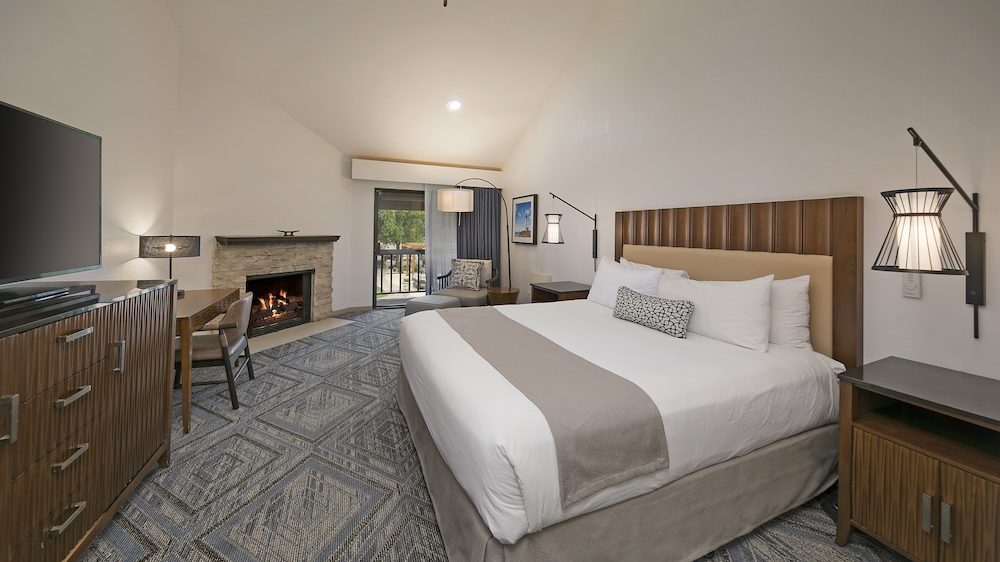 King-sized bed with white linens and a beige throw blanket in a modern hotel room. The room features a wooden headboard, two bedside tables with wall-mounted lamps, a patterned carpet, a flat-screen TV on a wooden dresser, a desk with a chair, a stone fireplace, and a seating area with a chair and floor lamp near a sliding glass door leading to a balcony. The walls are white, and the ceiling is vaulted with a ceiling fan.