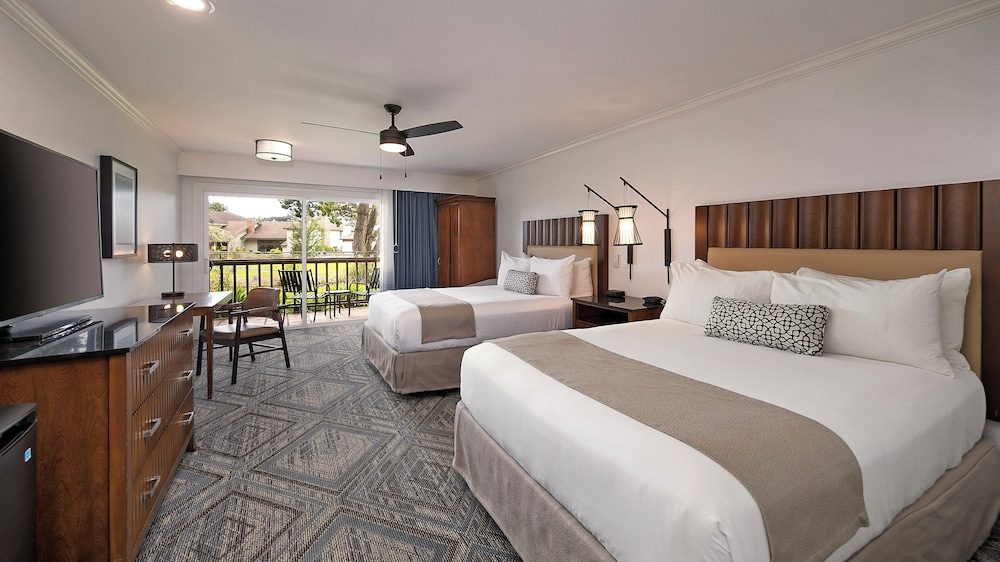 A modern hotel room with two beds featuring white linens, beige runners, and patterned accent pillows. The room has a geometric-patterned carpet, a wooden dresser with a flat-screen TV, a small desk with a chair, and a ceiling fan with lights. There is a sliding glass door leading to a balcony with outdoor seating and a view of greenery and neighboring buildings. The walls are light-colored, and the room includes wall-mounted lamps beside the beds.