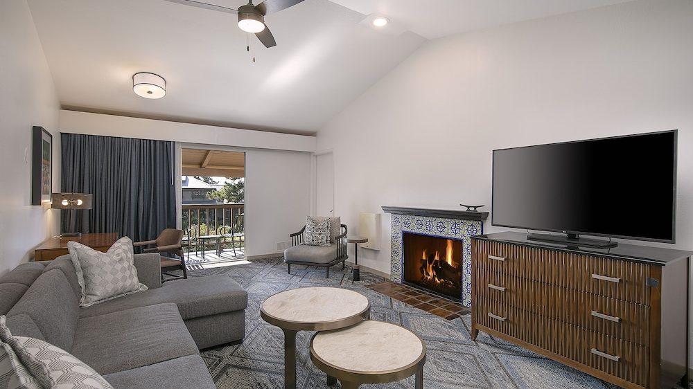 Living room with a gray sectional sofa, two round marble-top coffee tables, a patterned area rug, a fireplace with decorative tile surround, a flat-screen TV on a wooden cabinet, and a sliding glass door leading to a balcony with outdoor seating.
