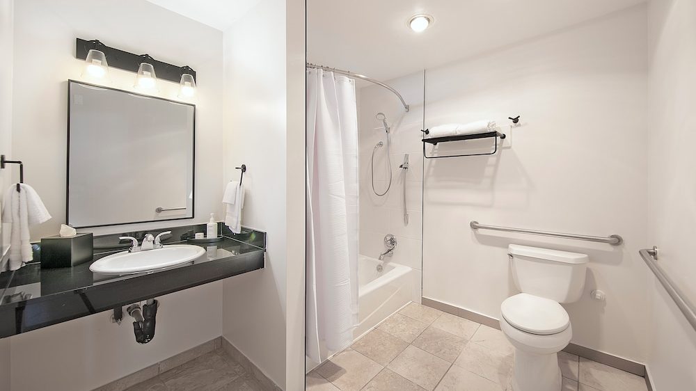 Bathroom with a white bathtub and shower, a white toilet, and a black countertop with a white sink. The walls are white, and the floor is tiled in a light beige color. There is a large mirror above the sink with three light fixtures, and towel racks with white towels are mounted on the walls. The bathroom has grab bars near the toilet and bathtub for accessibility.
