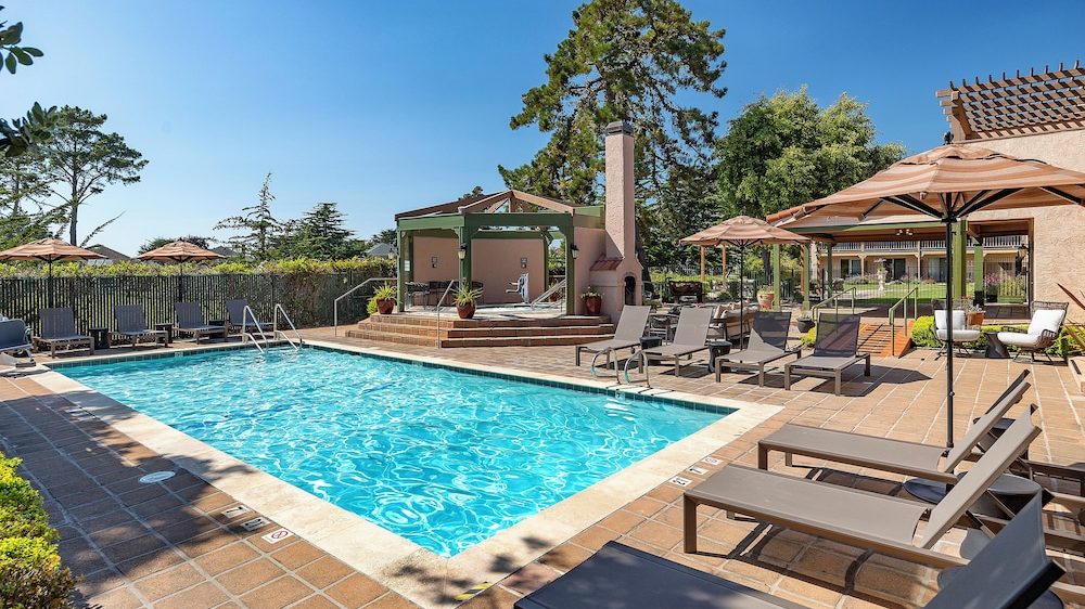 Outdoor swimming pool with clear blue water surrounded by tiled deck. Multiple lounge chairs and umbrellas are arranged around the pool area. A small covered patio with a fireplace and potted plants is visible in the background, along with trees and a clear blue sky.