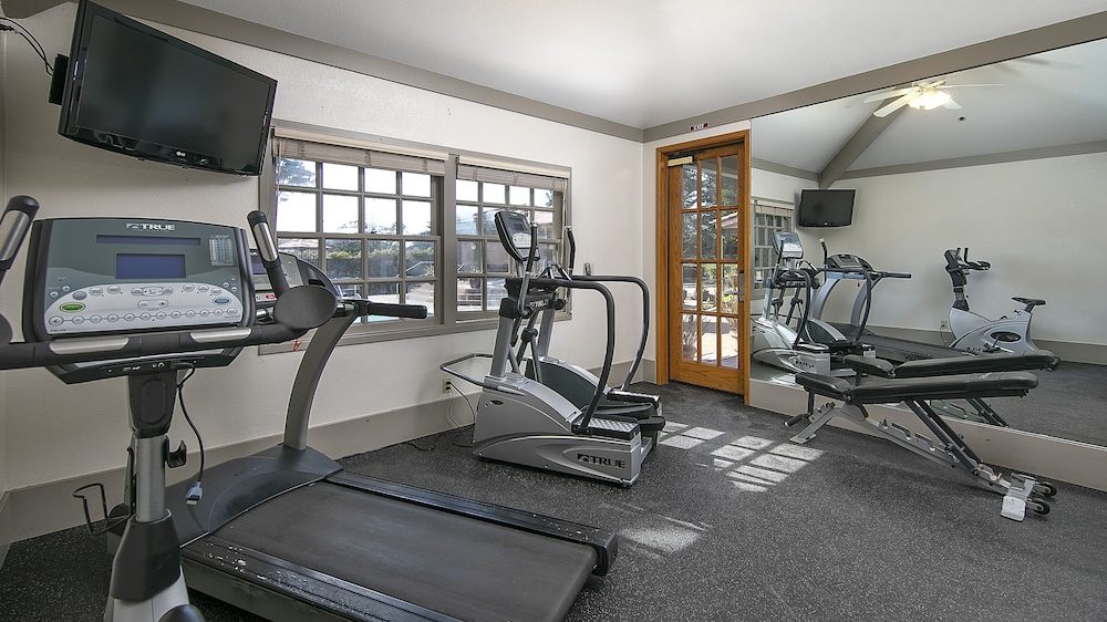 A small gym room with exercise equipment including a treadmill, an elliptical machine, a stationary bike, and a weight bench. There is a large wall mirror reflecting the equipment, a wall-mounted TV above the treadmill, and a window letting in natural light. The floor is covered with dark rubber gym flooring.