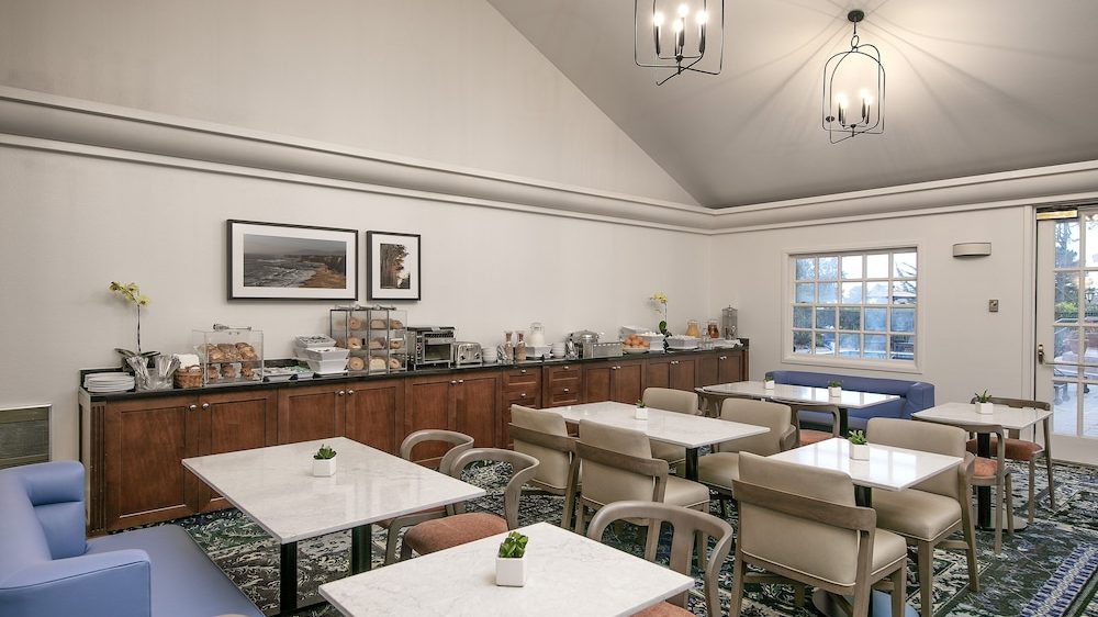 A dining area with multiple white marble-top tables and beige cushioned chairs arranged neatly on a patterned carpet. Along one wall, there is a long wooden cabinet with various breakfast items including pastries, a toaster, and beverage dispensers. The room has high vaulted ceilings with two modern black metal pendant lights and large windows letting in natural light. There are also two framed landscape photographs on the wall above the cabinet.