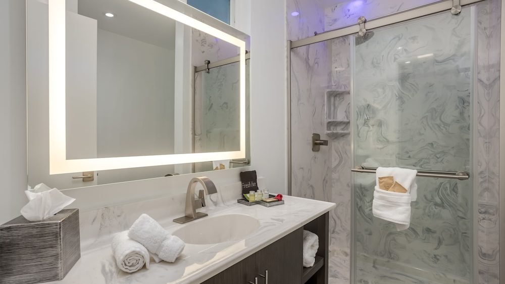 Modern bathroom with a marble countertop featuring a built-in sink, a large illuminated rectangular mirror, and a tissue box. The shower has sliding glass doors with a towel hanging on the handle, and the walls have a marble-like pattern. The space is well-lit with recessed lighting.