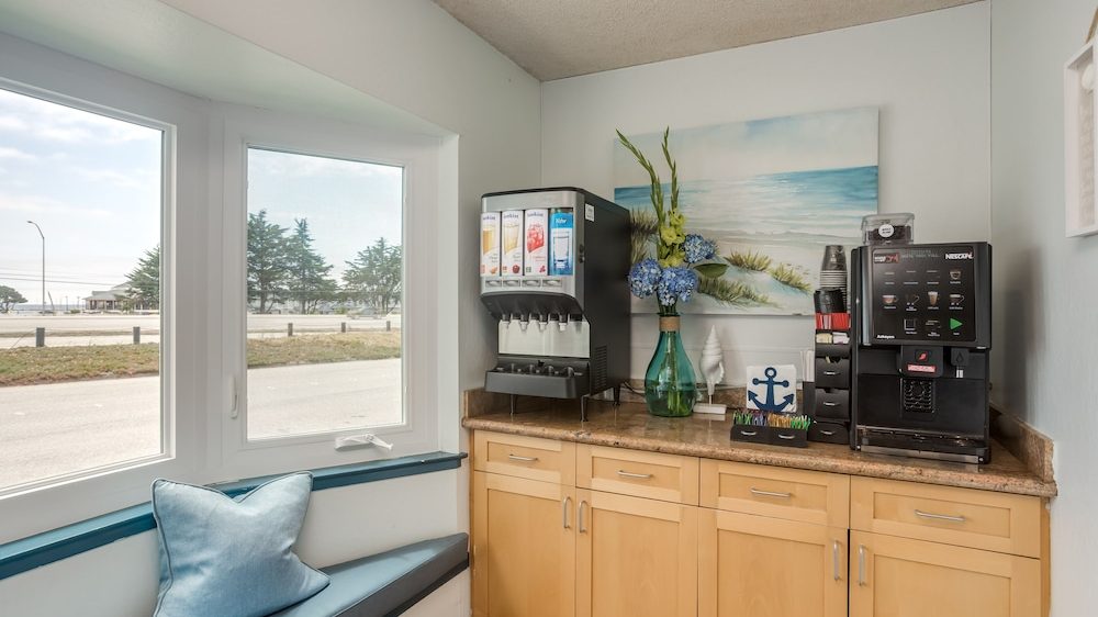 Countertop with a beverage dispenser offering various drink options, a Nescafé coffee machine, and a small organizer holding cups and stirrers. A green vase with blue and yellow flowers sits between the machines. Above the counter is a painting of a beach scene. Below the counter are light wood cabinets. To the left, a window seat with a blue cushion and pillow is visible.