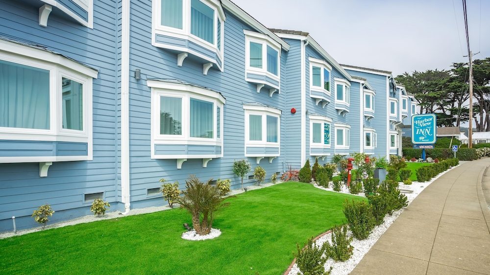 A blue two-story building with white-trimmed bay windows and a well-maintained green lawn in front. There are small bushes and plants along the edge of the lawn, bordered by white gravel. A sidewalk runs alongside the building, and a blue sign reading "Harbor View Inn" is visible near the entrance.