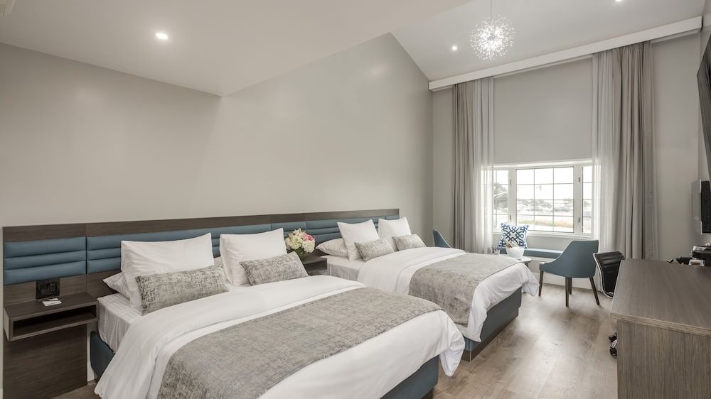 Two neatly made beds with white linens and gray patterned throws in a modern hotel room. The room features a wooden floor, a large window with sheer and blackout curtains, a small seating area with two blue chairs and a table, and a flat-screen TV mounted on the wall opposite the beds. The walls are painted light gray, and a contemporary light fixture hangs from the ceiling.
