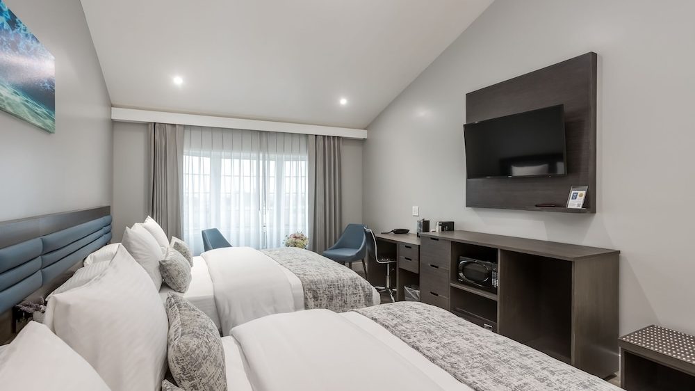 Two neatly made beds with white linens and gray patterned throws in a modern hotel room. The room features a wall-mounted flat-screen TV above a dark wood cabinet with a microwave, a desk with a chair, and two blue accent chairs near a large window with sheer and gray curtains. The walls are painted light gray, and the ceiling is vaulted with recessed lighting.