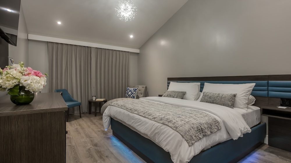 Modern bedroom with a large bed featuring a blue upholstered base and headboard, white bedding, and gray textured throw and pillows. The room has light wood flooring, gray walls, and a ceiling with recessed lighting and a decorative pendant light. A wooden dresser with a vase of flowers is on the left, and a small seating area with a blue chair, a gray armchair, and a round wooden table is near the window with gray curtains. Blue LED lighting is visible under the bed.