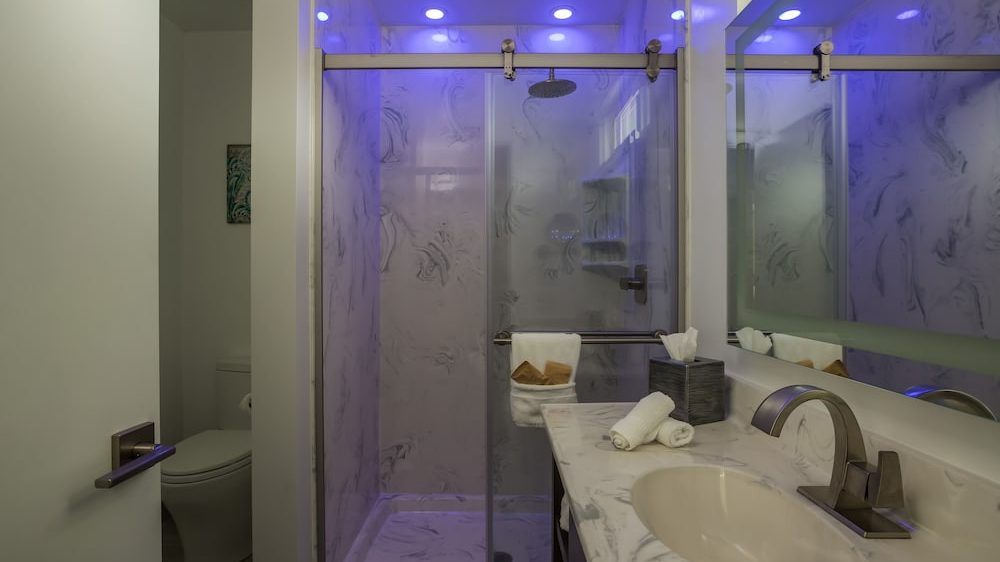Modern bathroom with a marble countertop sink, a tissue box, and rolled white towels. The shower has frosted glass sliding doors with a stainless steel frame and a rain showerhead. Blue LED lights illuminate the shower area. A toilet is partially visible behind a slightly open door.