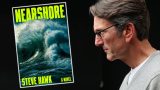 A man with gray hair and glasses, wearing a light gray sweater over a white shirt, is looking intently at a book cover titled "NEARSHORE" by Steve Hawk. The book cover features a large, powerful ocean wave with dark blue and green tones, and the title and author's name are in bold, bright yellow letters. The background behind the man is black.