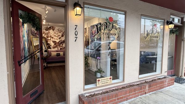 Art By You Studio storefront with beige walls and two large windows displaying the studio's logo. The entrance door is open, painted red with a wreath decoration. Inside, a purple couch is visible against a wall with large floral artwork. A small sign in the window reads "Sometimes Open, Sometimes Closed, Mostly Open." The address number 707 is displayed next to the door, and two wall-mounted lantern-style lights illuminate the entrance.