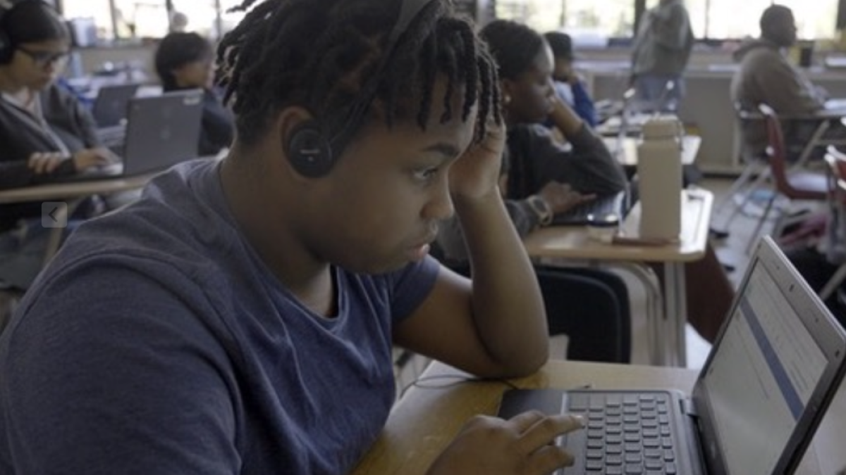 A student wearing headphones and a blue shirt is focused on typing on a laptop in a classroom. Other students in the background are also working on laptops, and a teacher stands near the windows. The classroom has large windows letting in natural light.