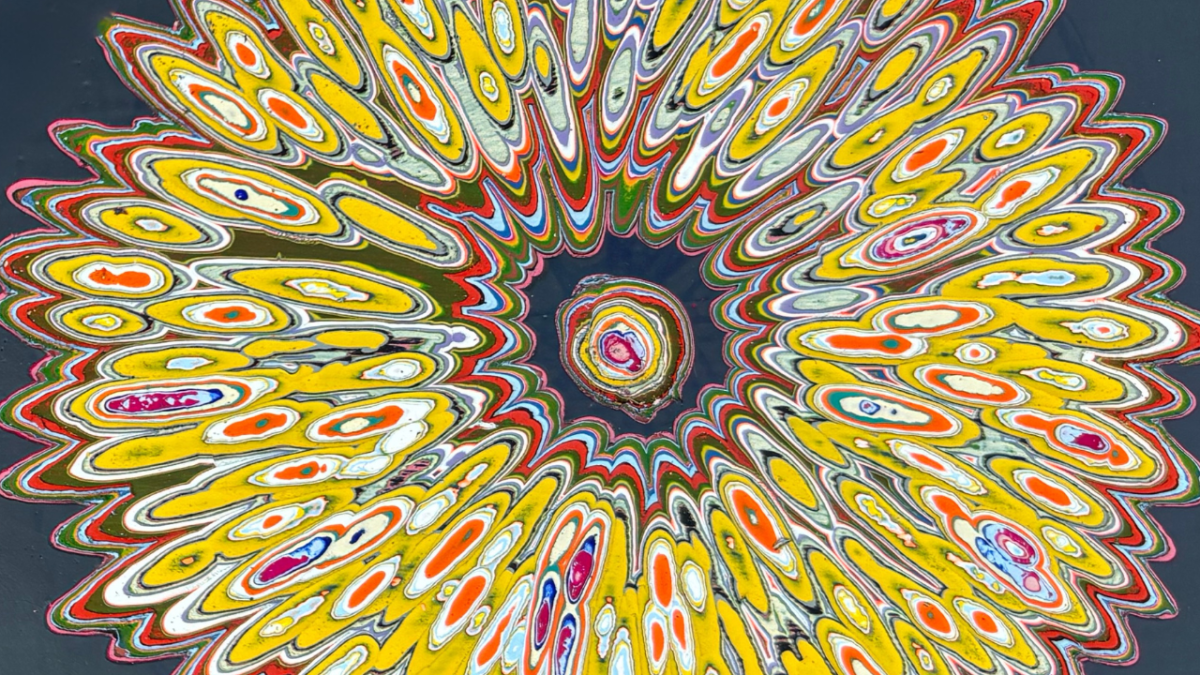 A vibrant, abstract circular pattern with concentric, irregular oval shapes radiating outward. The design features bright yellow, orange, white, red, blue, and green colors layered in a fluid, marbled style against a dark background. The center has a small, multicolored swirl, and the overall effect resembles a dynamic, colorful burst or flower.