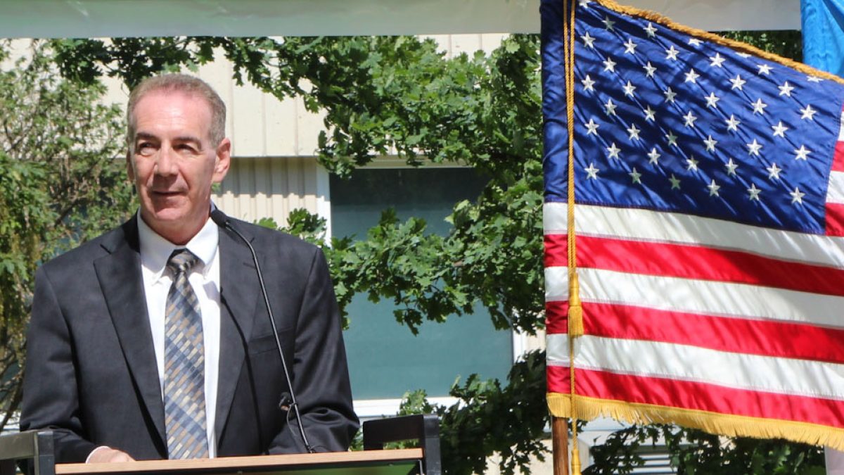 Man in a suit speaking at a podium outdoors beside a large American flag.