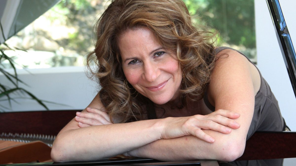 A woman with curly, light brown hair is leaning on a grand piano with her arms crossed. She is smiling gently and wearing a sleeveless dark top. The background includes a window with greenery outside.