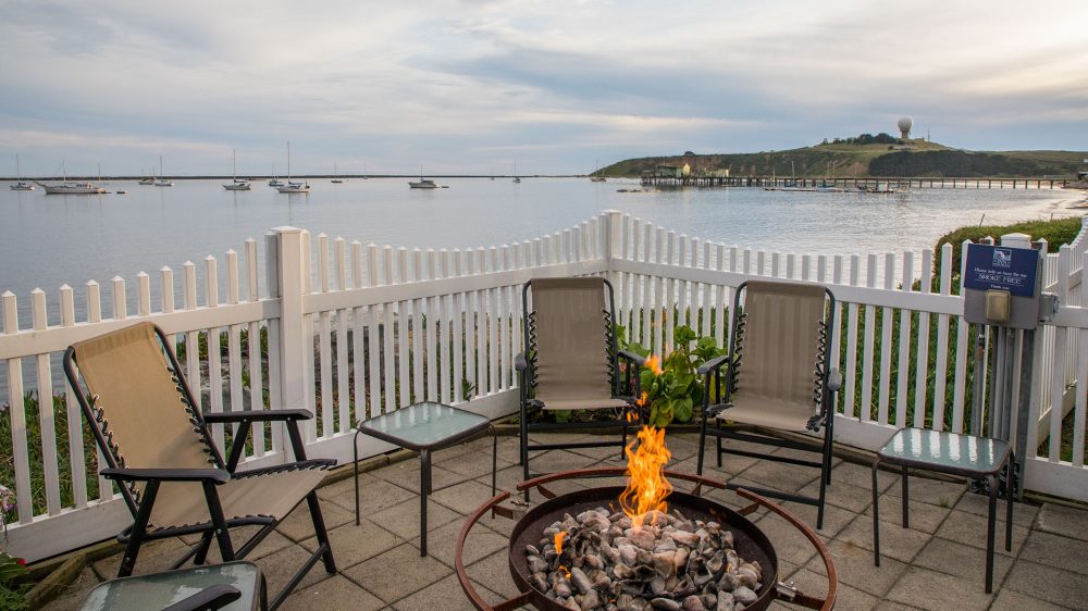 A patio area with four beige mesh chairs and two small glass-top tables arranged around a circular fire pit with flames. The patio is enclosed by a white picket fence, overlooking a calm bay with several sailboats anchored. In the background, there is a grassy hill with a large spherical radar dome and a pier extending into the water. The sky is partly cloudy.