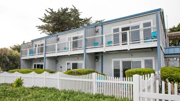 Two-story gray building with white trim and blue accents, featuring sliding glass doors and a balcony with white railing on the upper floor. Blue chairs are placed on the balcony and patio areas. A white picket fence and green bushes surround the building, with tall trees in the background.