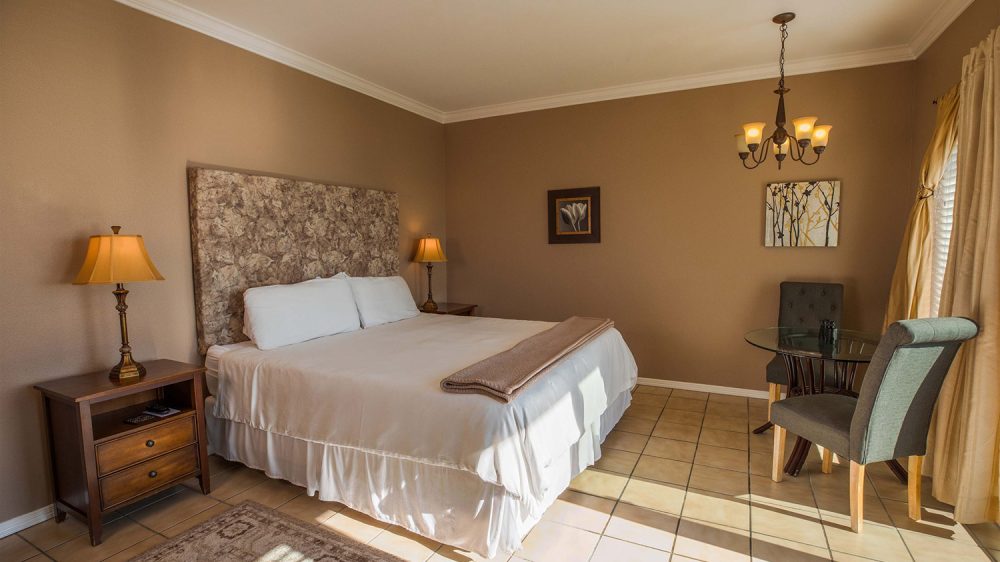 Bedroom with a large bed featuring a patterned headboard, white bedding, and a folded beige blanket. Two wooden nightstands with lamps flank the bed. The room has beige walls, tiled floor, a beige rug, and a glass-top table with two upholstered chairs near a window with beige curtains. A chandelier hangs from the ceiling, and two framed artworks are on the walls. Sunlight streams through the window, casting shadows on the floor.