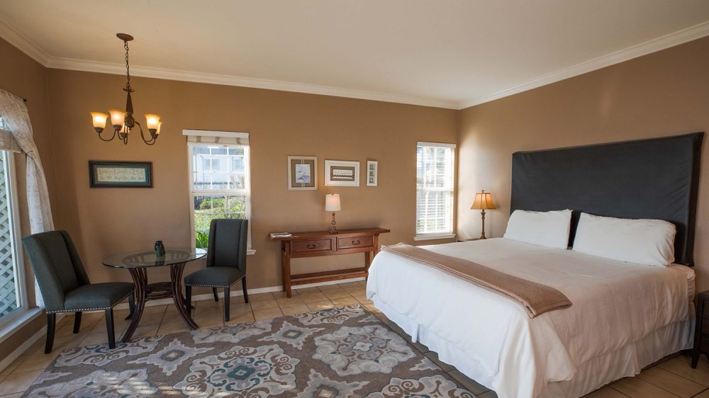 Bedroom with beige walls and tiled floor, featuring a large bed with white bedding and a dark headboard. A patterned area rug covers part of the floor. Two windows with white blinds allow natural light in. A wooden console table with a lamp and framed pictures hangs on the wall between the windows. A small round glass table with two dark upholstered chairs is positioned near the left window. A chandelier with five lights hangs from the ceiling.