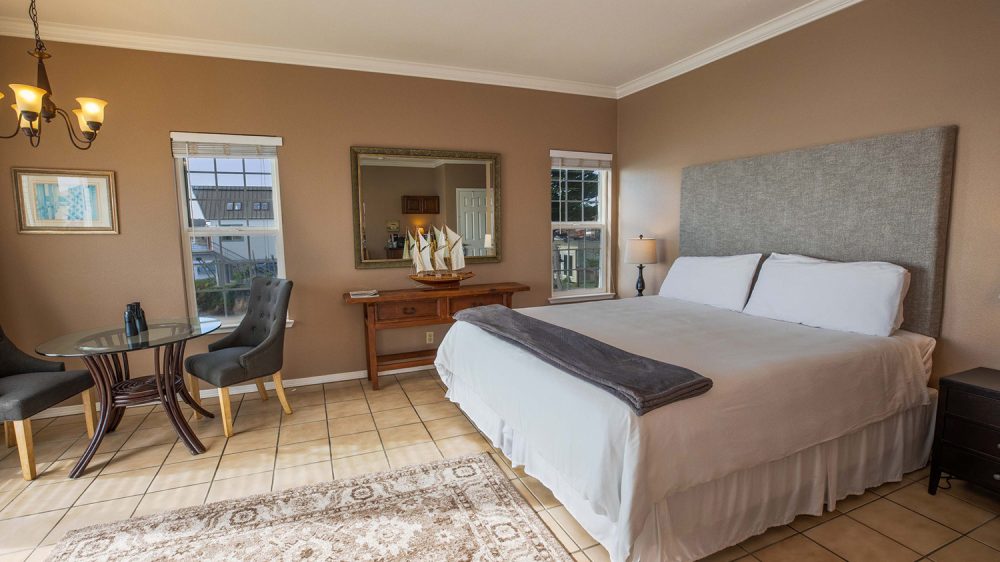 Bedroom with a large bed featuring a gray upholstered headboard, white bedding, and a folded gray blanket at the foot. The room has beige walls, tiled floors, and a patterned area rug. There is a wooden console table with a decorative sailboat model and a large framed mirror above it. Two windows with white blinds flank the mirror. A small round glass table with two gray upholstered chairs is positioned near one window. A dark nightstand with a lamp is beside the bed. A chandelier with three lights hangs from the ceiling.
