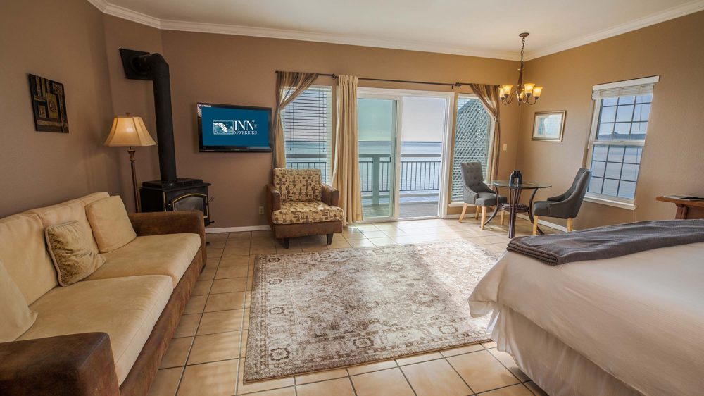 Room with beige and brown tones featuring a beige sofa with brown armrests, a patterned armchair, a glass-top round table with two gray upholstered chairs, a bed with white linens and a gray blanket, a floor lamp, a wall-mounted TV displaying "Inn at Mavericks," a wood stove with a black chimney pipe, a large area rug, tiled floor, and sliding glass doors leading to a balcony overlooking water.