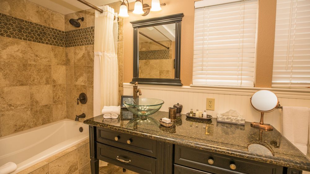 Bathroom with a dark granite countertop vanity featuring a glass vessel sink and bronze faucet, a black-framed mirror, and a bathtub with beige tile surround accented by a hexagonal tile border. The countertop holds neatly folded white towels, toiletries, a tissue box, and a small round mirror. White blinds cover the windows above the vanity.