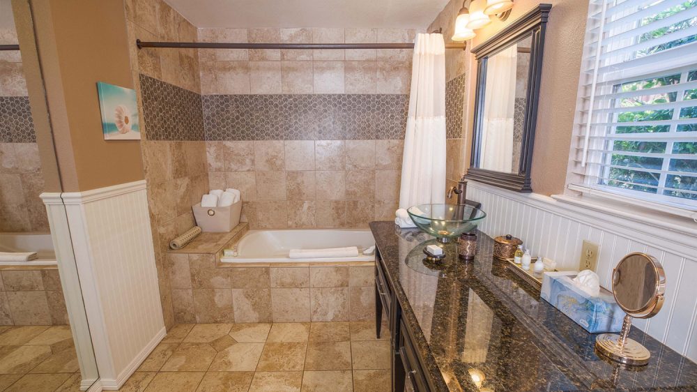 Bathroom with beige tiled walls and floor, featuring a built-in bathtub with a white shower curtain. A basket with rolled white towels sits on the bathtub ledge. The vanity has a dark granite countertop with a glass vessel sink, bronze faucet, and a large framed mirror above. On the countertop are toiletries, a tissue box, a small round mirror, and decorative containers. A window with white blinds lets in natural light.
