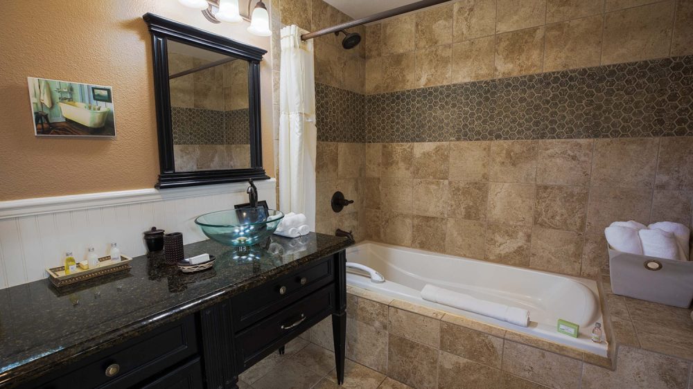 Bathroom with a black granite countertop and a glass vessel sink on a dark wooden vanity. Above the sink is a black-framed mirror with three light fixtures. The bathtub is surrounded by beige and brown tiled walls, featuring a horizontal strip of hexagonal mosaic tiles. A white shower curtain hangs on a curved rod, and white towels are neatly rolled and placed on the tub edge and in a gray basket on a tiled ledge. A small framed picture of a bathtub hangs on the wall above the vanity.