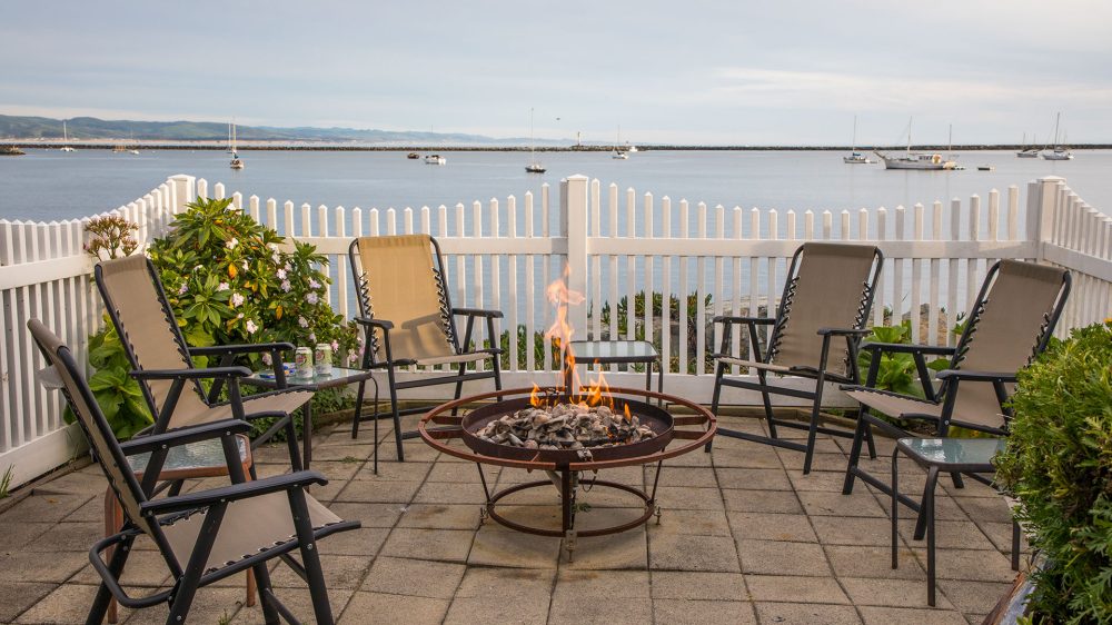 Outdoor patio area with six beige sling chairs arranged around a round fire pit with flames, set on a tiled floor. The space is enclosed by a white picket fence, overlooking a calm body of water with several sailboats anchored in the distance. Small glass-top side tables are placed between some of the chairs, and there are green bushes and flowering plants along the fence.