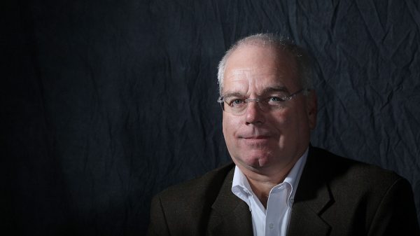 A middle-aged man with short gray hair and glasses, wearing a dark blazer over a white collared shirt, is set against a dark, textured background. He has a neutral expression and is looking slightly to the side.